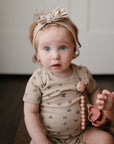 A light-skinned young child with curly reddish hair and blue eyes, wearing a beige floral onesie and headband, sits indoors holding a toy and the mushie Silicone Pacifier Clip | Hera, gazing curiously at the camera.