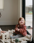Two young children play on a blanket by a large window. One smiles in a floral top, the other looks at the camera. The mushie Silicone Pacifier Clip | Hera adds minimalist style as light filters gently from outside.