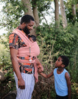 A man outdoors, dressed in a patterned shirt and striped pants, carries a baby in a pink wrap with the mushie Silicone Pacifier Clip | Eva. He holds hands with a boy in a blue tank top, surrounded by lush greenery.
