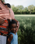 A person stands in a field, carrying a baby in a pink wrap with the FRIGG Daisy Silicone Pacifier 2-Pack clipped on. Nearby, a young child in a blue shirt looks at the camera, surrounded by trees and greenery.