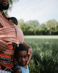 A man in a patterned shirt holds a baby in a pink sling with the mushie Silicone Pacifier Clip | Eva attached. A young child stands nearby, leaning into his arm, with green fields and trees in the background.