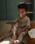 A young child plays barefoot and focused in a cozy, softly lit room, using the FRIGG Daisy Natural Rubber Pacifier 2-Pack with its daisy-shaped design and natural rubber latex nipple.