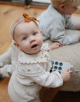 A baby wearing a brown bow headband holds the mushie Phone Press Toy, looking up beside an older child playing on a cream cushion. They’re indoors on a wooden floor, enjoying sensory play together.