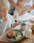 A young blonde child enjoys a meal with pastry and fruit from a divided plate, while a baby sits nearby. The wooden table is protected by a mushie Silicone Placemat, along with matching cups and plates.