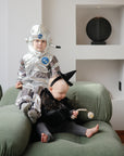 Two young children sit on a green couch—one in a shiny astronaut costume, the other in a sparkly witch dress holding a FRIGG Daisy Natural Rubber Pacifier by FRIGG and a beaded toy. Decor items are displayed on shelves in the background.