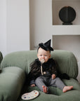 A baby in a black sparkly witch costume and pointed hat, using the mushie Silicone Pacifier Clip | Hera, sits on a green couch near a paint palette, with white shelves and round decor enhancing the room’s minimalist style.