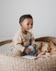 A baby in a beige knit outfit sits in a woven basket with a polka-dot cushion, holding a blue toy and using the mushie Silicone Pacifier Clip | Eva to secure their pacifier. A tan blanket is placed beside the baby.