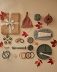 A flat lay of baby items on a beige background with autumn leaves, featuring mushie's Snack Cup, a knotted hat, folded blanket, teether, toys, divided plate, and a tube of cream.