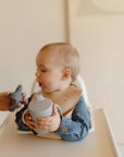 A baby in a high chair holds the Mushie Trainer Sippy Cup with easy-grip handles, gazing at an adult offering a gray silicone pacifier. The baby wears a beige bib and denim shirt against a softly lit, neutral background.