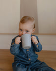 A young toddler in denim sits on a wooden floor, holding and drinking from a Mushie Trainer Sippy Cup with both hands, gazing upward with wide blue eyes.
