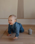 A smiling baby in a denim outfit crawls on a wooden floor with one hand reaching forward. Nearby, a Mushie Trainer Sippy Cup with easy-grip handles rests on the floor.