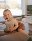 A smiling baby in a brown outfit sits on a couch, wearing a beige mushie Muslin Bib and holding a gray toy block. Sunlight streams through the window, with a round coffee table in the background.
