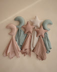 Four mushie Star Lovey Blankets—each with a plush moon or star top and pastel organic cotton cloths (two dusty pink moons, one white star, one sage green moon)—are arranged on a beige background.