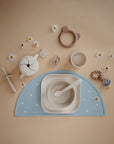 A flat lay of mushie Square Dinnerware Plates in beige, BPA-free plastic, with a bowl, spoon, cup, sippy cup, pacifiers, teether, and flower-shaped toys arranged around a blue placemat with small white flowers.