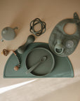 A set of sage green baby feeding essentials—including a mushie Silicone Pacifier Clip | Eva—plus plate with spoon, sippy cup, teether, bib, bead toy, and placemat, all made from food-grade silicone and neatly arranged on a light surface.