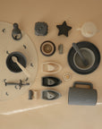 A flat lay of mushie neutral-toned baby feeding and teething items—food-grade silicone bowls, cups, spoons, teethers, boats—and a mushie Silicone Placemat with animal illustrations, all arranged on a beige background.