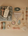 Flat lay of baby care essentials on beige, featuring a towel, lotion bottle, rattles, soft mitten, teething rings, handled cup, pacifiers with food-grade silicone accessories, and the mushie Silicone Pacifier Clip | Eva in gentle pastel hues.