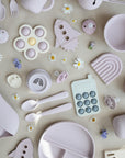 The mushie Flower Press Toy, along with pastel-colored silicone baby utensils and toys such as spoons, plates, a rocket, a rainbow, and a toy phone, is arranged on a neutral background with scattered white and purple flowers.