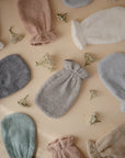 Mushie Organic Cotton Bath Mitts, soft and textured in pastel colors, are arranged on a beige surface with small white flowers. Cozy and plush, they're ideal for bathing or self-care and perfect for sensitive skin.