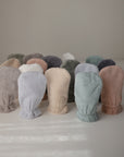 A selection of Mushie Organic Cotton Bath Mitts, ideal for sensitive skin, is displayed upright on a white surface in neutral and pastel shades, highlighting their soft, textured fabric.