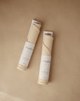 Two rolled-up mushie Silicone Placemats made from food-grade silicone, in minimalist “mushie” packaging, rest on a light brown surface at slight angles to each other.