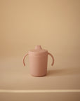 A pink Mushie Trainer Sippy Cup with easy-grip handles and a spout lid sits on a beige surface with a matching background. The non-toxic cup features the embossed "mushie" logo on its side.