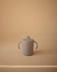 A Mushie Trainer Sippy Cup in gray with easy-grip handles and a spout is centered on a beige background. The non-toxic cup, featuring "mushie" on its side, highlights Mushie's minimal, soft design.