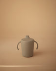 A gray Mushie Trainer Sippy Cup with easy-grip handles and a secure lid is centered on a beige background. The silicone cup, designed for toddlers, features the Mushie brand name embossed on its surface and is made from non-toxic materials.