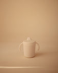 The Mushie Trainer Sippy Cup, featuring easy-grip handles and a spout lid, rests on a smooth beige surface with a matching background. This simple, non-toxic cup is designed for little hands and perfect for everyday use.