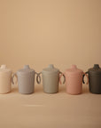 Five Mushie Trainer Sippy Cups with easy-grip handles in white, light blue, sage green, pink, and dark gray are arranged in a row on a beige background. Each Mushie Trainer Sippy Cup is non-toxic and designed for little hands.