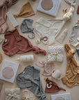A flat lay of mushie baby essentials in neutral and pastel hues, featuring the Ribbed Baby Bonnet, onesies, bibs, bottles, pacifiers, boxes, and wrapped gifts, all artfully arranged on a soft beige background.