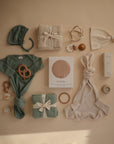 Flat lay of neutral and green baby items on beige, including clothing, blankets, teething toys, a mushie Ribbed Baby Bonnet, headband, bunny ear lovey, and a boxed pacifier holder.