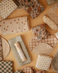 The mushie Organic Cotton Muslin Burp Cloth 2-Pack, featuring floral, checkered, polka dot, and paisley patterns, is neatly arranged on a peach-colored background.