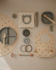 The mushie Silicone Pacifier Clip | Eva, made from food-grade silicone, is shown with a bib, divided plate, spoon, bowl, cup, teether rings, and a polka-dotted cloth napkin on a beige surface in neutral tones.