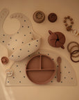A flat lay of baby and toddler feeding items in neutral tones, featuring a patterned bib, divided plate, spoon, silicone placemat, pacifier, teething rings, wooden beads, and the mushie Snack Cup with a spill-proof design.