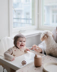 A smiling baby in a knitted bonnet sits in a high chair as an adult woman feeds them with a mushie Silicone Feeding Spoon. Sunlight streams through large windows, and the woman holds a small white dog.