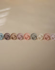 Nine FRIGG Andersen Fairytale Silicone Pacifier 2-Packs by FRIGG, in pastel shades, are neatly lined up on a beige surface. Their scalloped edges reveal a soft gradient from blue to pink, softly illuminated by natural light from the left.