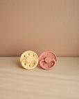 The FRIGG Moon Silicone Pacifier 2-Pack features one pale yellow and one muted pink pacifier, both with medical grade silicone nipples for comfort and safety, resting side by side on a light beige surface.