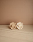 The Mushie FRIGG Rope Natural Rubber Pacifier 2-Pack features two beige pacifiers with round handles and textured edges, resting side by side on a neutral surface for soothing comfort and a seamless look.