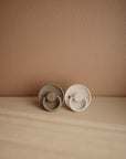 Two beige FRIGG Rope Silicone Pacifiers from the FRIGG 2-Pack are displayed side by side on a smooth surface against a light brown wall, with textured edges and natural lighting creating a warm, calming atmosphere for baby.