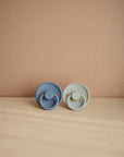 The Mushie FRIGG Rope Natural Rubber Pacifier 2-Pack features blue and light gray pacifiers with twisted rope-like edges, displayed side by side on a beige surface against a peach wall.