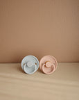The Mushie FRIGG Rope Natural Rubber Pacifier 2-Pack, in light blue and light pink, is displayed side by side on a beige surface against a light brown wall. Each pacifier features a comfortable natural rubber latex nipple.