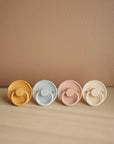 The Mushie FRIGG Rope Natural Rubber Pacifier 2-Pack features pastel yellow, blue, pink, and cream pacifiers with natural rubber latex nipples and textured rims—perfect baby essentials displayed against a tan background.