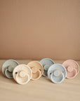 Six FRIGG Rope Silicone Pacifier 2-Packs by FRIGG, made from medical grade silicone, feature round handles and come in soft cream, light blue, and pale pink shades. Arranged neatly on a neutral surface against a beige background.
