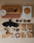 A flat lay of baby items—clothes, teething toys, a bat plush, hat, swaddle, star night light, boxed muslin crib sheet, and the mushie Silicone Pacifier Clip | Eva—all in soft neutral and earthy tones.