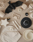 Flat lay of mushie baby products in cream and black, featuring silicone teethers, rattles, spoons, bowls, pacifiers, baby oil, a muslin wrap, an Organic Cotton Muslin Burp Cloth 2-Pack by mushie, and a glittery black hat with a bat toy.