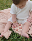 A baby in a white outfit sits on grass atop a peach blanket, wearing bracelets and exploring the mushie Flower Press Toy—an engaging beige, paw-shaped sensory toy.