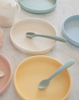 Mushie Silicone Feeding Spoons, shown with pastel silicone plates and a white sippy cup on marble, offer safe, stylish feeding for kids. Made from food-grade silicone, they're perfect for your child’s meals.