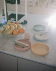 A marble countertop displays a mushie Silicone Feeding Spoon on each of three pastel plates, alongside a pink silicone block, fruit bowl with bananas and oranges, and white candles in bright sunlight.
