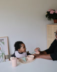 An adult feeds a smiling baby in a high chair with a bowl, utensils, and the Mushie Trainer Sippy Cup with easy-grip handles. Colorful art and flowers brighten the scene as they share this joyful mealtime.